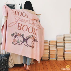 pink booksters gonna book hooded blanket for readers in front of a pile of books organized on the floor