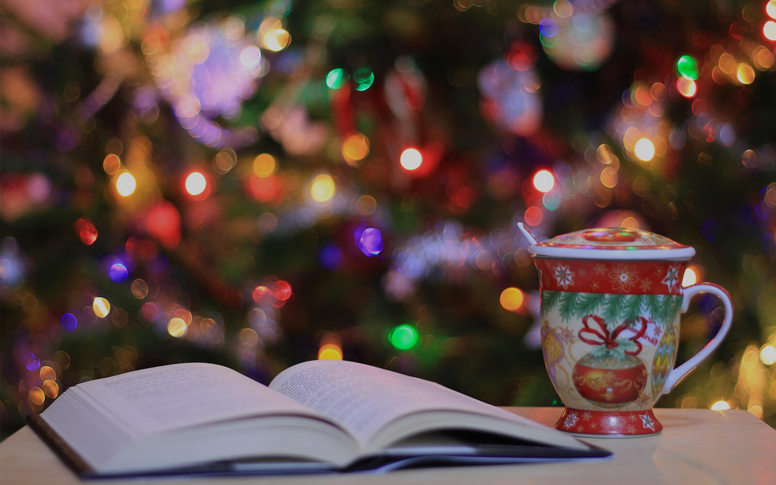 an open book next to a holiday themed mug and in the background there's a christmas tree decorated with lights